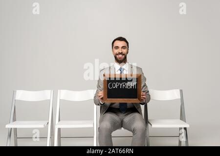 Lächelnder Geschäftsmann, der mit Wort-Copyright auf dem Stuhl auf grauem Hintergrund sitzt Stockfoto