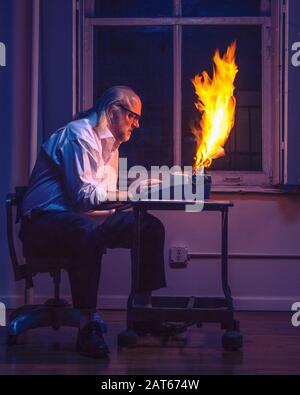 Ein kahler Mann mittleren Alters, der auf einer alten Schreibmaschine tippt, ist nachts vor einem dunklen Fenster in Flammen aufgegangen. Stockfoto