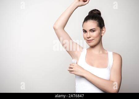 Eine positive Frau, die die Kamera betrachtet, während sie Deodorant auf den Achselhöhenkerchen anwendet, isoliert auf Grau Stockfoto
