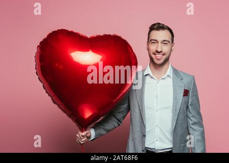 Bärtiger lächelnder Mann mit rotem Herzballon, isoliert auf Rosa Stockfoto