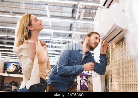 Freund schaut auf Klimaanlage und lächelnde Freundin, die im Haushaltsgerätehaus kühlt Stockfoto