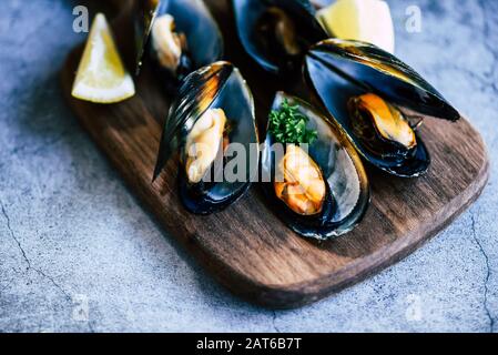 Gekochte Muscheln mit Kräutern Zitrone und dunklem Plattenhintergrund / Frische Meeresfrüchte Schalentiere auf Holzschneidebrett im Restaurant Muschelschalennahrung Stockfoto