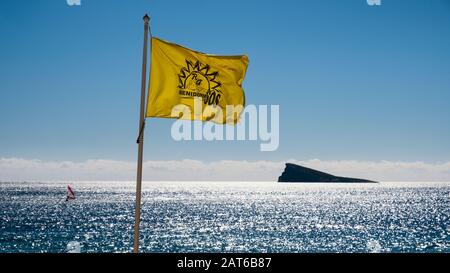 Schwenkende gelbe Flagge des Rettungsschwimmers mit der Insel Benidorm im Hintergrund an einem sonnigen Tag mit blauem Himmel (Benidorm, Marina Baixa, Costa Blanca, Alicante, Spanien) Stockfoto