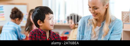 Panoramaaufnahme von lächelnder Lehrerin und Kind mit Kindern im Hintergrund in der montessori-schule Stockfoto