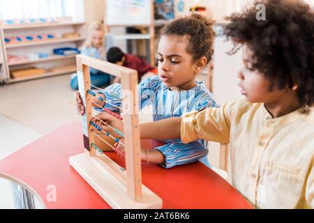 Selektiver Fokus von afroamerikanischen Kindern, die mit Noten mit Lehrer und Kind im Hintergrund in der montessori-schule spielen Stockfoto