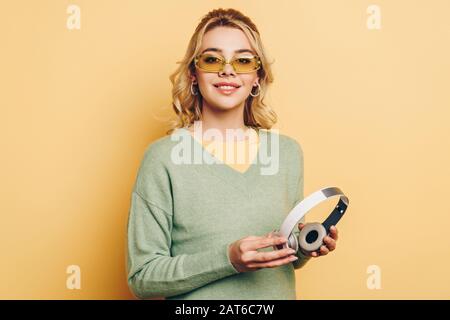 Hübsches Mädchen in einer Brille, die drahtlose Kopfhörer hält und mit der Kamera auf gelbem Hintergrund lächelt Stockfoto