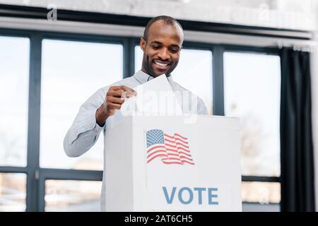 Glücklicher afroamerikanischer Mann, der mit der Flagge amerikas abstimmen und Wahlzettel in die Box legen wird Stockfoto