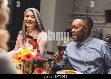 Selektive Fokussierung der lächelnden asiatischen Frau und des afroamerikanischen Mannes, der während des Abendessens Weingläser hält Stockfoto