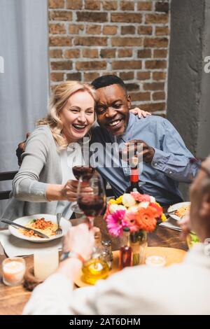 Selektive Fokussierung von lächelnden multikulturellen Freunden, die sich beim Abendessen umarmen und klinken Stockfoto