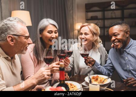 Lächelnde multikulturelle Freunde, die beim Abendessen mit Weingläsern sprechen und klinken Stockfoto