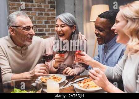 Lächelnde multikulturelle Freunde, die beim Abendessen mit Weingläsern sprechen und klinken Stockfoto