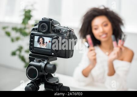 Selektiver Fokus der Digitalkamera mit lockig afroamerikanischem Videoblogger in Klammern, die Lippenglosses auf dem Display halten Stockfoto