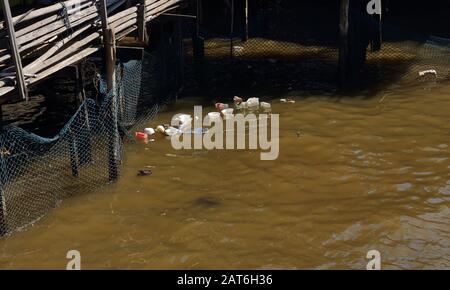 Plastikflaschen und Schaumstoff-Lebensmittelbehälter, die im Fluss in der Nähe des Netzes weggeworfen werden, gebunden an Holzkonstruktion am Wasser, Klimaschutzkonzept Stockfoto