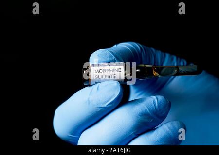 Morphinsulfat-Injektionsampulle, die von der Handschuhhand des Gesundheitsperformers mit dunklem Hintergrund gehalten wird. Stockfoto