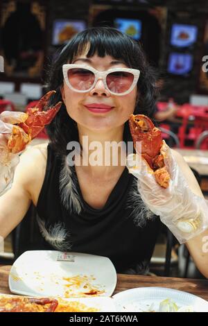 Eine chinesin mittleren Alters in Sonnenbrille und Plastikhandschuhen, die Singapur-Chili-Krabben an einem Esstisch im Lebensmittel-Stall essen Stockfoto