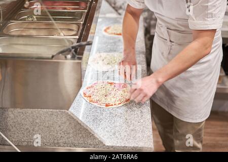 Pizza kochen. Ordnet Käsezutaten auf der Teigvorform an. Nahaufnahme der Küchenchef-Bäckerei in einer einheitlichen weißen Schürze in der Küche Stockfoto