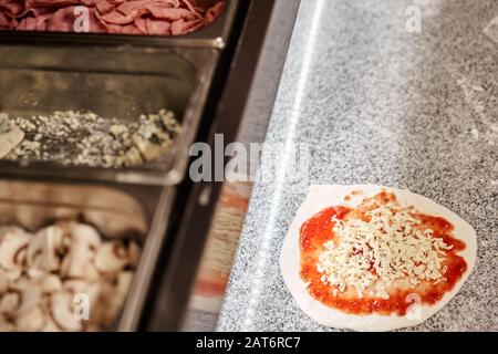 Pizza kochen. Ordnet Käsezutaten auf der Teigvorform an. Nahaufnahme der Küchenchef-Bäckerei in einer einheitlichen weißen Schürze in der Küche Stockfoto