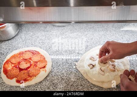 Pizza kochen. Legt Champignons und Sahnesauce aus, Zutaten auf der Teigvorform. Nahaufnahme der Küchenchef-Bäckerei in einer einheitlichen weißen Schürze in der Küche Stockfoto