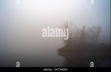 Teich während des kalten Herbstmorgens im Nebel bedeckt. Dichter Nebel und Reflexionen in ruhiger Wasseroberfläche. Stockfoto