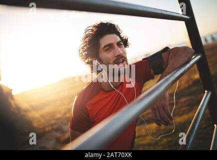 Portrait eines jungen männlichen Athleten, der Musik auf seinen Ohrhörern hört und sich nach dem laufen am sonnigen Tag eine Pause gönnt Stockfoto