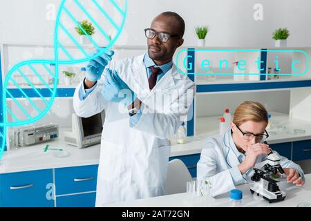 afroamerikanischer Biologe hält Reagenzglas und steht in der Nähe von dna-Illustrationen, Kollege mit Mikroskop Stockfoto