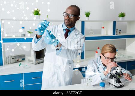 Der afroamerikanische Biologe, der das Reagenzglas hält und in der Nähe der Illustration steht, sein Kollege mit dem Mikroskop Stockfoto