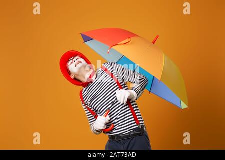 MIME man, der mit mehrfarbigem Regenschirm in der Hand aufblickt, isoliert auf leerem orangefarbenem Hintergrund im Studio Stockfoto