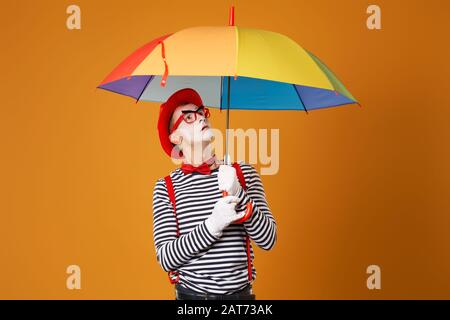 MIME-Suche mit mehrfarbigem Regenschirm in der Hand isoliert auf leerem orangefarbenem Hintergrund im Studio Stockfoto