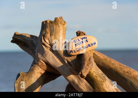 "Alles, was Sie brauchen, ist Aldeburgh", geschrieben auf Stein, der auf etwas Treibholz am Aldeburgh Beach platziert ist Stockfoto