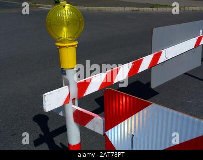 Straßenbarriere aus rot-weißen Holzbohlen auf Kunststoffpfosten mit bernsteinfarbener mobiler Sicherheit oder Gefahrenlicht. Grauer Asphaltstraßenbelag. Stockfoto
