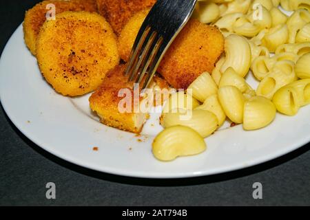 Ein Stück Nuggets auf einer Gabel und ein weißer Teller mit gebratenen Hühnerhuhn Nuggets, Pasta und Ketchup auf dunklem Hintergrund. Nahaufnahme Stockfoto