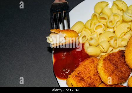 Ein Stück Nuggets auf einer Gabel und ein weißer Teller mit gebratenen Hühnerhuhn Nuggets, Pasta und Ketchup auf dunklem Hintergrund. Nahaufnahme Stockfoto