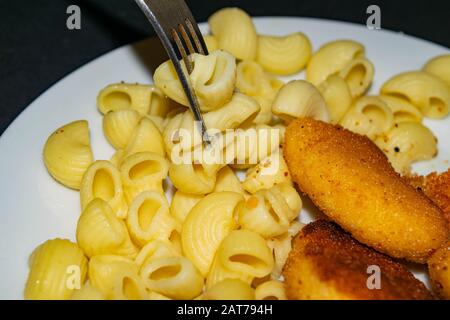 Ein paar gekochte Makaroni auf einer Gabel und eine weiße Platte mit gebratenen Hühnerhuhn Nuggets, Makkaroni und Ketchup auf dunklem Hintergrund. Nahaufnahme Stockfoto