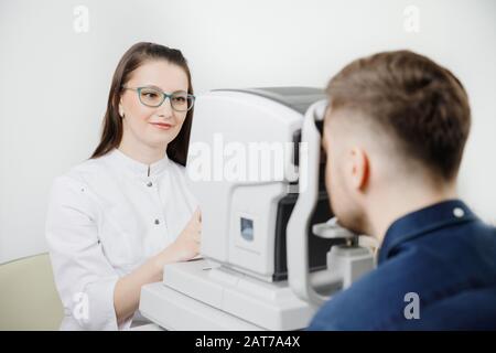 Attraktive Frau Arzt Augenarzt mit grün-randbewohnter Brille überprüft die Sicht des jungen Mannes auf dunkle Haare und blaues Hemd in der Moderne Stockfoto