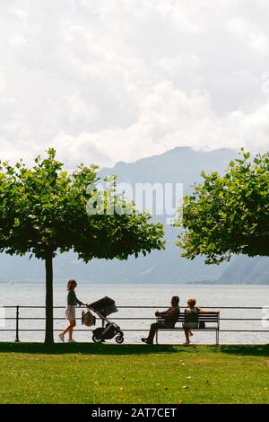 Eine junge Frau mit Kinderwagen und einem pensionierten Paar auf einer Bank mit Blick auf den Vierwaldstättersee in Brunnen, Schwyz, Schweiz EU Stockfoto