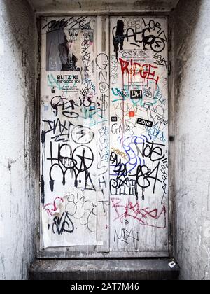 Urbane Anarchie. Detail einer versenkten weißen Tür, die mit Graffiti-Tags bedeckt ist, die ein deformiertes Warnschild abwehrt. Stockfoto