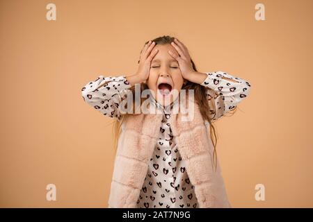 Süßes kleines Mädchen schließt ihre Ohren mit der Hand und schreit auf beigem Hintergrund. Stockfoto