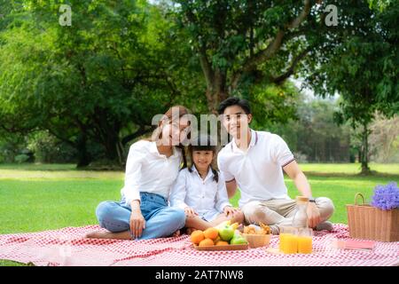 Asiatische Teenager Familie fröhlicher Urlaub Picknick-Moment im Park mit Vater, Mutter und Tochter Blick auf Kamera und Lächeln für einen glücklichen Urlaub zu verbringen Stockfoto