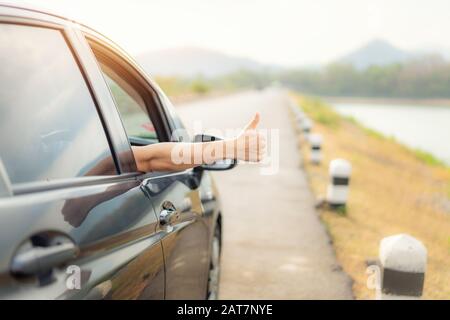 Hand des Reisenden mit Daumen nach oben aus dem Auto aus der Asphaltstraße gehen Sie mit wunderschönem Landschaftssee und Bergen und Sonnenlicht am Abend auf Reisen. C Stockfoto