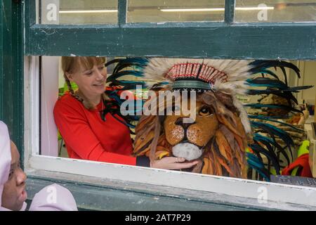 Weibliche Kunstlehrerin, die einen handgefertigten Löwenkopf aus Pappmaché durch ein offenes Fenster zeigt; Higgins Gallery, Bedford, Großbritannien Stockfoto