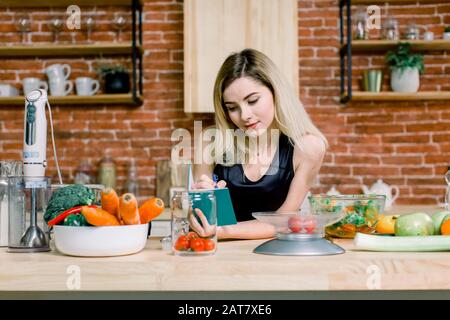 Fröhliche schöne blonde Frau, die in ihrer Küche steht und zu Hause eine Einkaufsliste auf einem Notizbuch schreibt. Gesunde Ernährung - Gemüsesalat. Ernährung. Nähren Stockfoto