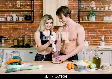 Gut gebauter gutaussehender Mann steht am Tisch in der Küche. Er hilft seinem hübschen Mädchen, Smoothies aus gesundem Essen zu mischen. Glückliches Paar, gesunder Lebensstil Stockfoto