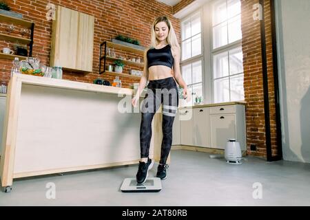 Dünne und schöne kaukasische blonde Frau, die in ihrer Küche auf Fußbodenwaage tritt. Abnehmen, Diät und gesunde Ernährung