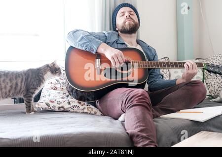 Verträumter Mann, mit geschlossenen Augen, spielt Gitarre, sitzt auf einem weichen Sofa, neben ihm ist eine Katze, die an dem Geschehen interessiert ist. Stockfoto
