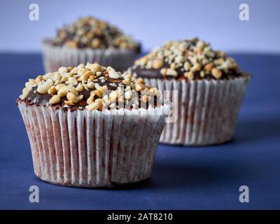 Drei Muffins mit Erdnüssen und Schokoladenglasur in Reihe auf blauem Hintergrund. Selektiver Fokus. Nahaufnahme. Stockfoto