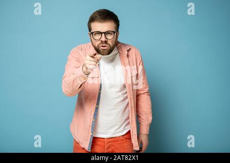 Der verärgerte kaukasische Mann zeigt mit dem Zeigefinger auf jemanden und gibt ihm die Schuld, indem er mit verengten Augen in die Kamera blickt. Stockfoto