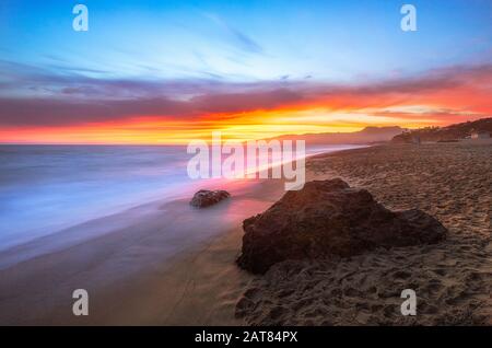 Das blaue, nebelige Wasser wird durch den langen Belichtungseffekt ergänzt. Er reflektiert den brennenden Sonnenuntergang mit orangefarbenen und roten Farbtönen und einem hellblauen Himmel. Stockfoto