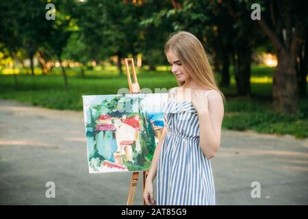 Schöne Mädchen-Künstlerin zeichnet im Park ein Bild mit einer Palette mit Farben und einem Spachtel. Staffelei und Leinwand mit einem Bild Stockfoto