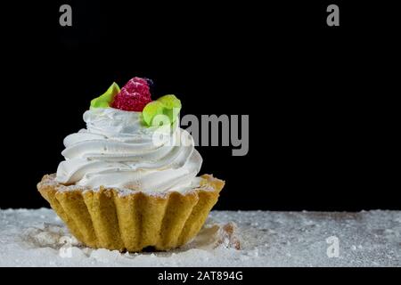 Kuchen mit weißer Creme, die mit Beeren auf einem Holzbrett auf schwarzem Grund verziert ist. Stockfoto