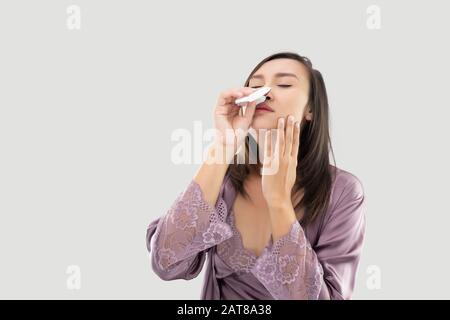 PM2.5 Stauballergie, asiatische Frauen in satinierter Nachtwäsche mit Nasenbluten vor grauem Hintergrund, asiatische Menschen atmen giftige Luft in der Wohnung Stockfoto